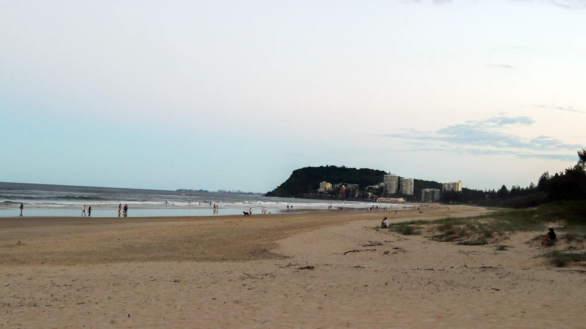 Burleigh Beach on the Gold Coast, featuring sandy shores, scattered visitors, a rocky headland in the distance, and nearby modern buildings.