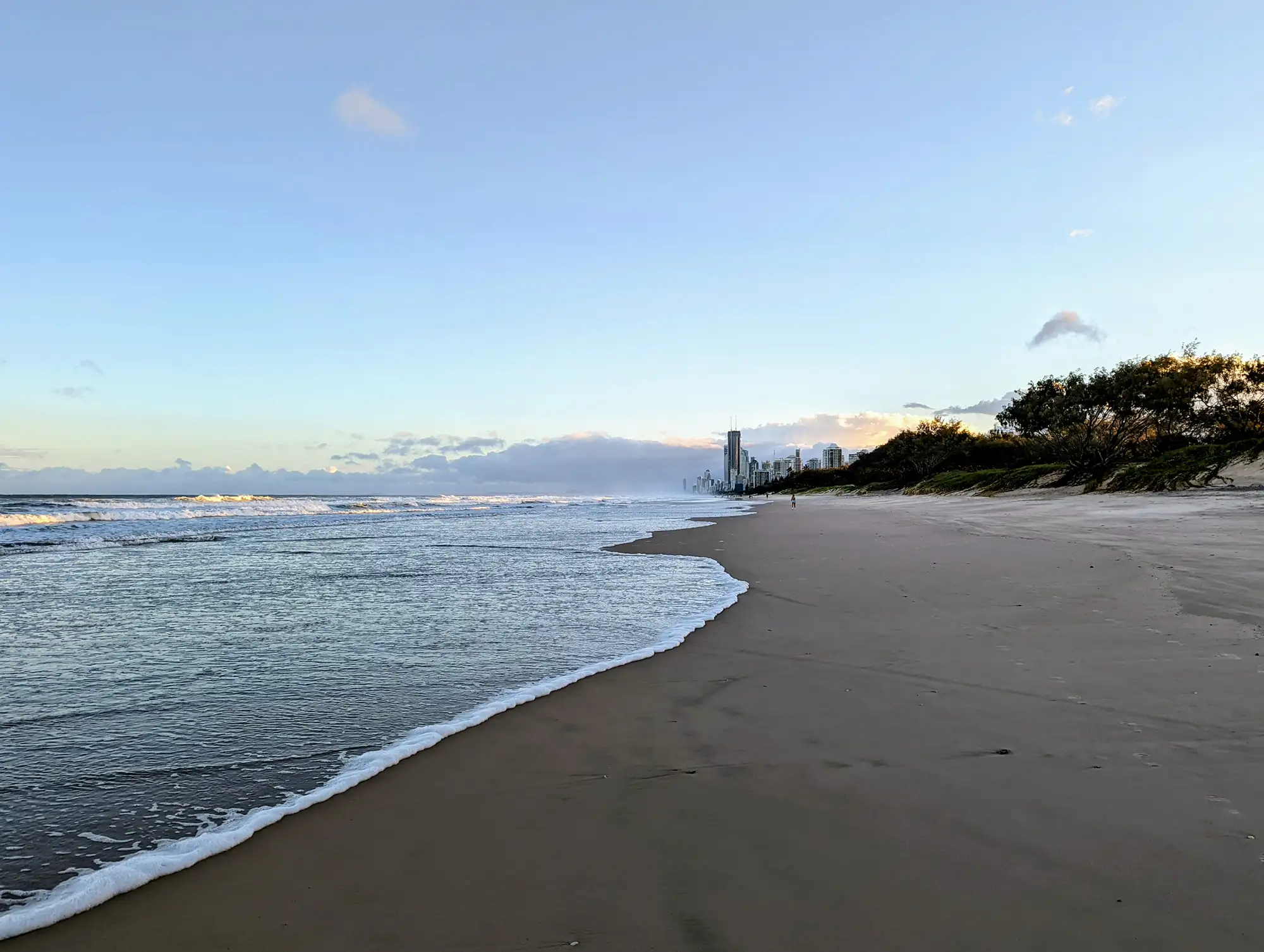 Wide sandy shoreline at The Spit Beach, Gold Coast, with gentle waves, distant skyscrapers, and greenery along the sand dunes.